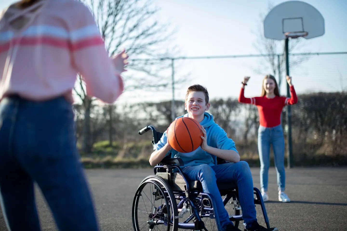 Ung gutt i rullestol med basketball, med ungdommer i bakgrunnen på basketballbane